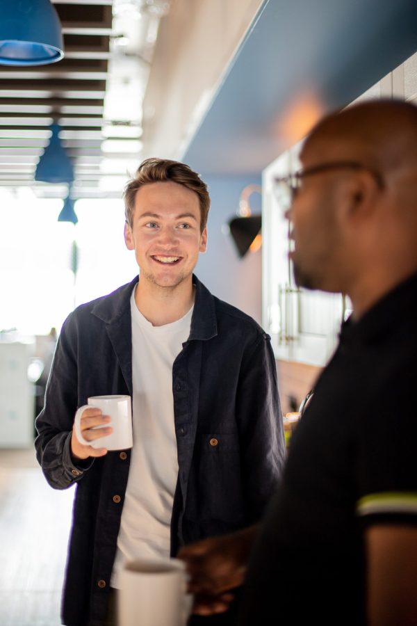 Two colleagues chatting and smiling while holding mugs in a bright office kitchen, symbolising supportive workplace relationships and mental health peer support