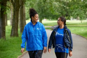 Two women walking together in a park, wearing blue Mind-branded jackets and t-shirts, promoting the Community Wellbeing Walk for Mental Health Awareness Week. Join us for a walk to support mental health and wellbeing during Mental Health Awareness Week. Get active, connect with others, and raise awareness for mental health support.