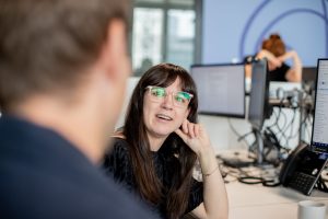 A woman with glasses engaging in a conversation in an office environment, promoting the 'Mental Health: Everything, Everywhere, Everyone – Building Stronger Communities at Work' webinar. Join us on Wednesday, 15th May 2025, from 12:00pm – 12:30pm for an insightful session on how fostering belonging, connection, and shared purpose can improve workplace mental health, enhance employee retention, and boost morale. Secure your spot for this essential 30-minute webinar.