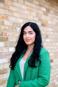 A woman with long, wavy black hair is standing in front of a light brick wall. She is a female volunteer for Mind in Mid Herts smiles confidently in front of a brick wall during Volunteers’ Week, celebrating mental health support and community service in Hertfordshire. She has a calm, confident expression and is looking directly at the camera.