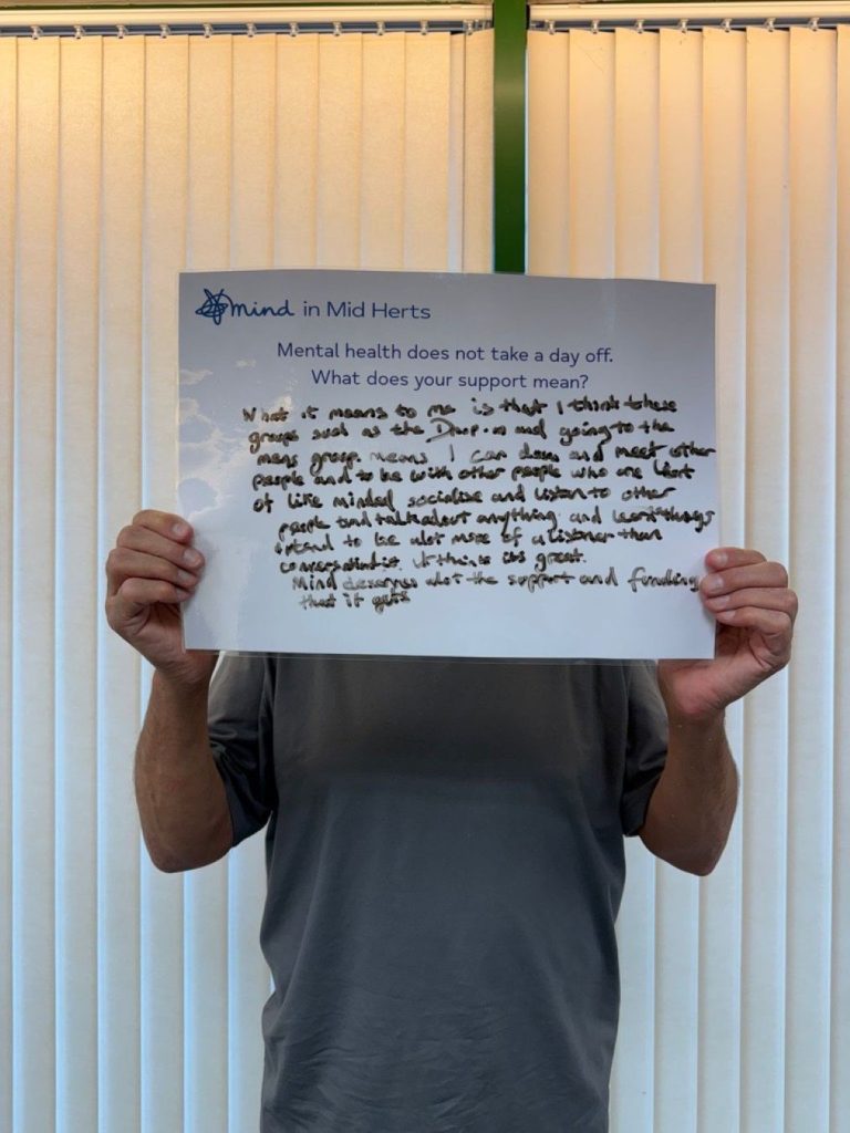 A person holding a handwritten sign that says: “Mind in Mid Herts. Mental health does not take a day off. What does your support mean? What it means to me is that I attend these groups such as the Drop-in and going to the men’s group means I can come and meet other people and be with other people who are in the same situation and understand one another. Mind deserves the support and funding that it gets.”.