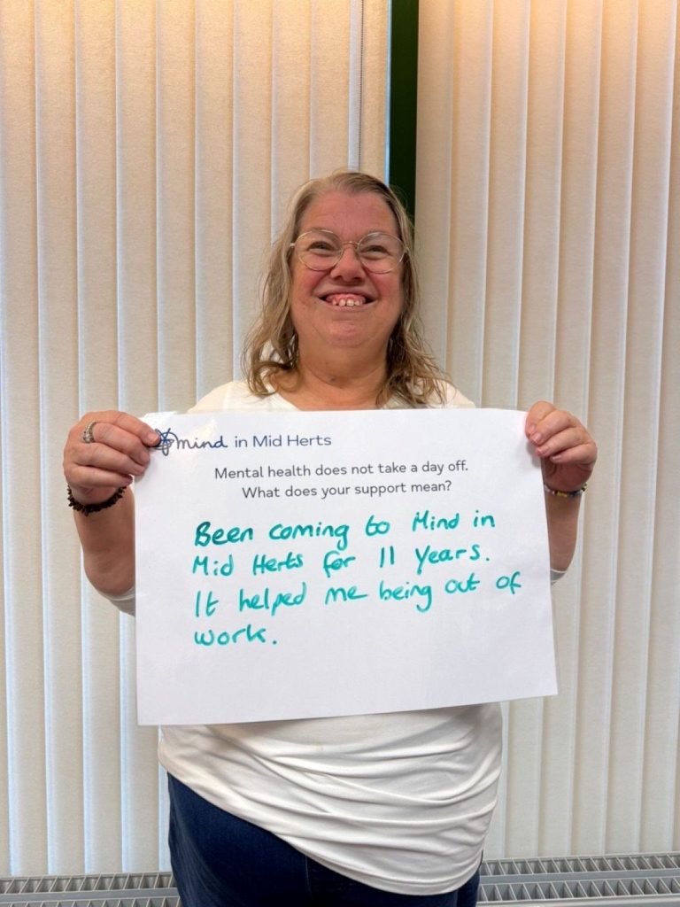 A smiling person wearing glasses and a white top holding a sign that reads: “Mind in Mid Herts. Mental health does not take a day off. What does your support mean? Been coming to Mind in Mid Herts for 11 years. It helped me being out of work.”