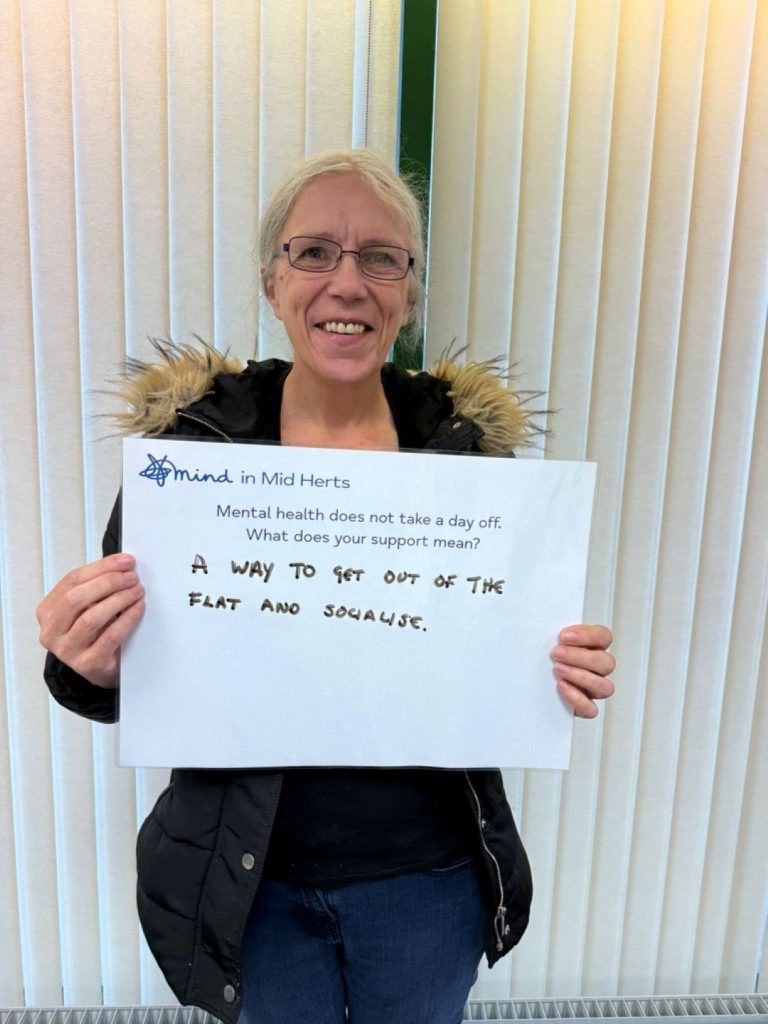 A smiling person wearing glasses and a black jacket with a fur-trimmed hood holds a sign reading: “Mind in Mid Herts. Mental health does not take a day off. What does your support mean? A way to get out of the flat and socialise.”