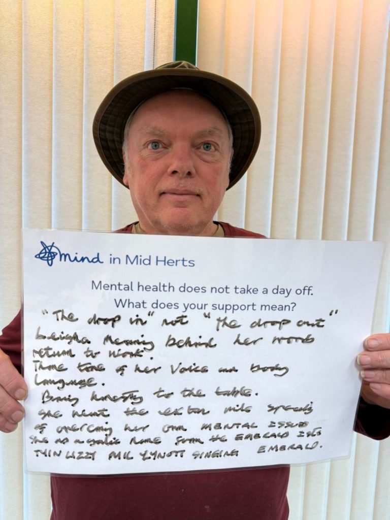 A person wearing a hat and maroon shirt holds a handwritten sign that says: “Mind in Mid Herts. Mental health does not take a day off. What does your support mean? ‘The drop in’, not ‘the drop out’. Bringing honesty to the table and supporting others through their mental health journeys.”