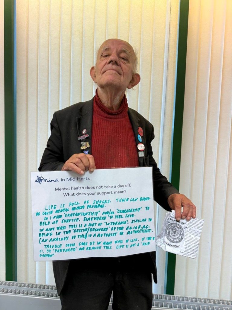A person in a red jumper and black jacket holds a handwritten sign that says: “Mind in Mid Herts. Mental health does not take a day off. What does your support mean? Life is full of shocks. There can be companionship and comradeship to help us survive. Mind helps people feel safe and supported.”