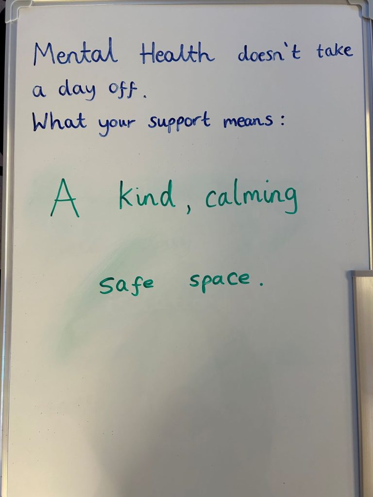 A close-up of a whiteboard with the handwritten message: “Mental health doesn’t take a day off. What your support means: A kind, calming safe space.”