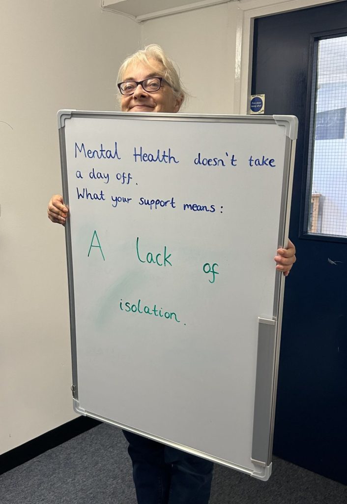 A person smiling and holding a whiteboard that says: “Mental health doesn’t take a day off. What your support means: A lack of isolation.”