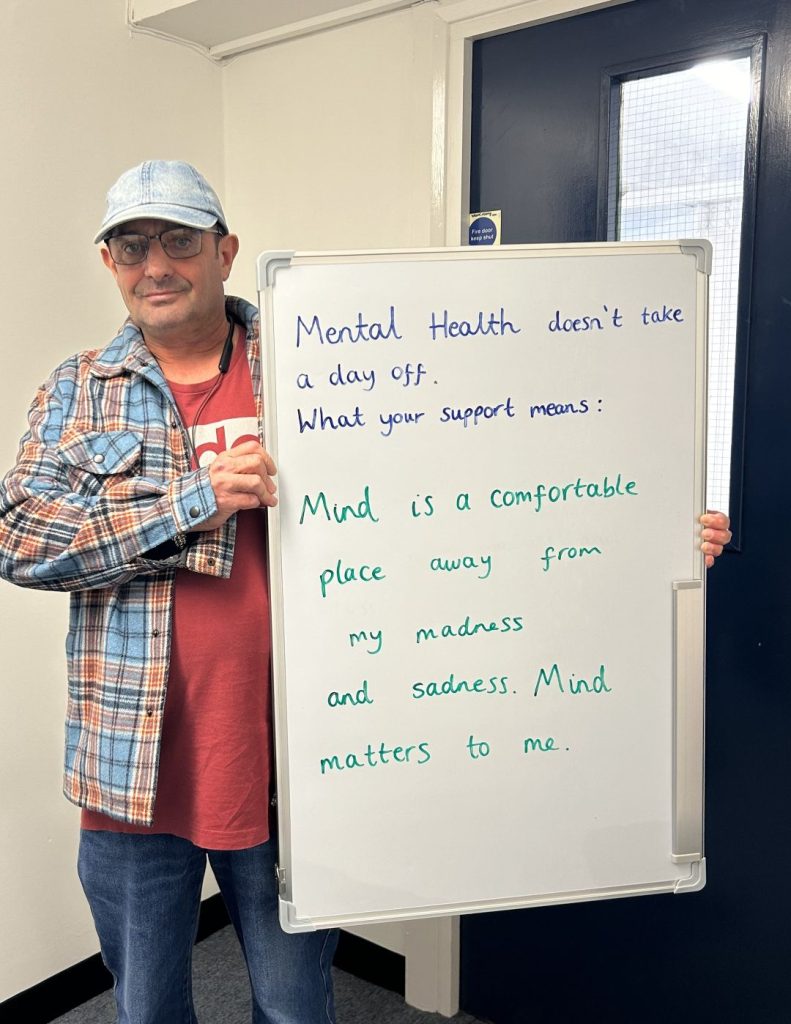 A person wearing glasses, a checked shirt and denim cap holds a whiteboard that reads: “Mental health doesn’t take a day off. What your support means: Mind is a comfortable place away from my madness and sadness. Mind matters to me.”