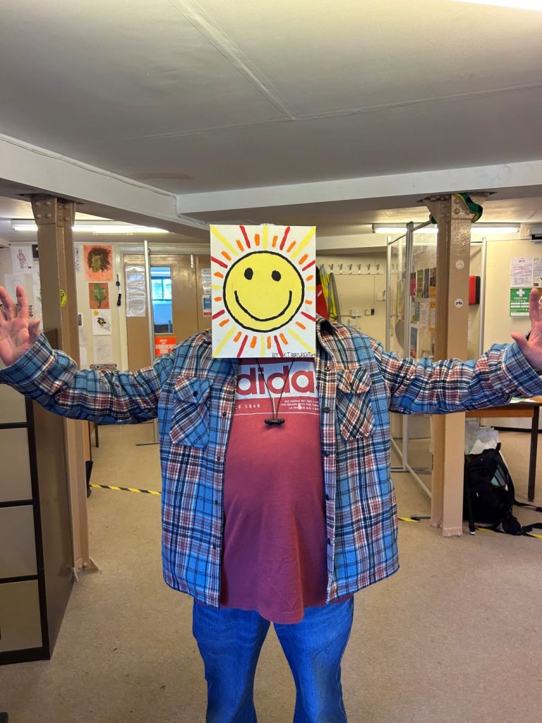 A person in a red shirt and checked shirt stands with arms open, holding a painting of a bright yellow smiling sun in front of their face.