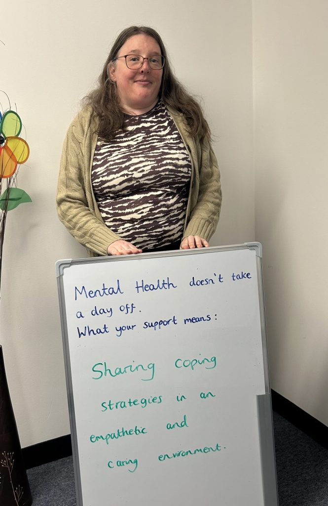 A person with glasses and long hair stands next to a whiteboard that says: “Mental health doesn’t take a day off. What your support means: Sharing coping strategies in an empathetic and caring environment.”