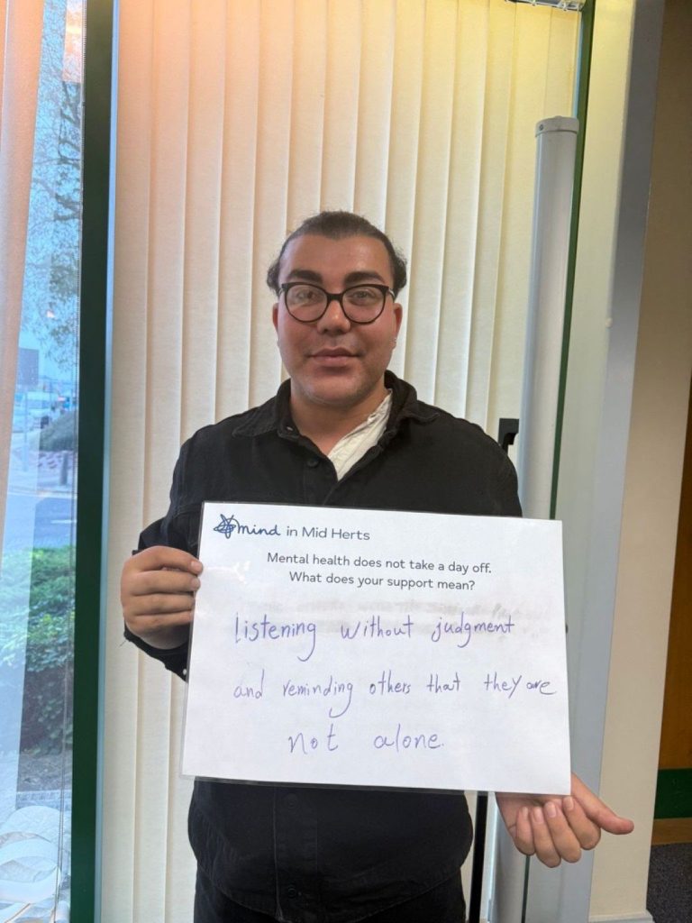 A person with glasses holding a sign that says: “Mind in Mid Herts. Mental health does not take a day off. What does your support mean? Listening without judgment and reminding others that they are not alone.”