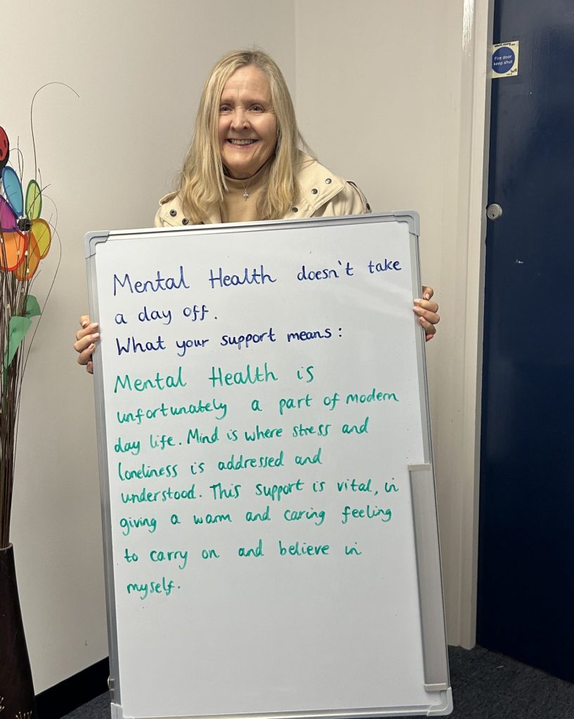 A smiling person in a light coat holds a whiteboard that reads: “Mental health doesn’t take a day off. What your support means: Mental health is unfortunately a part of modern life. Mind is where stress and loneliness are addressed and understood. This support is vital in giving a warm and caring feeling to carry on and believe in myself.”