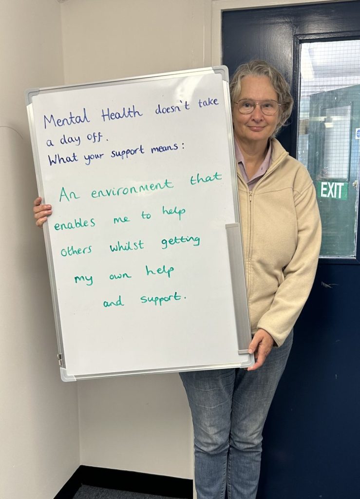 A person wearing glasses and a beige fleece jacket stands next to a whiteboard that reads: “Mental health doesn’t take a day off. What your support means: An environment that enables me to help others whilst getting my own help and support.”