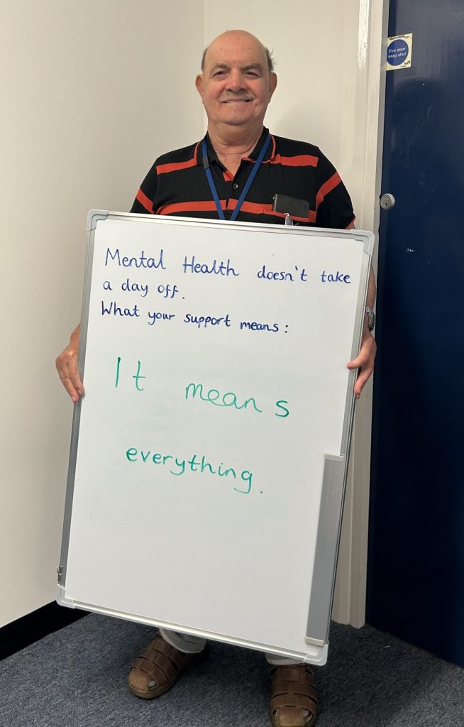 A smiling person in a black and red striped shirt holds a whiteboard that says: “Mental health doesn’t take a day off. What your support means: It means everything.”