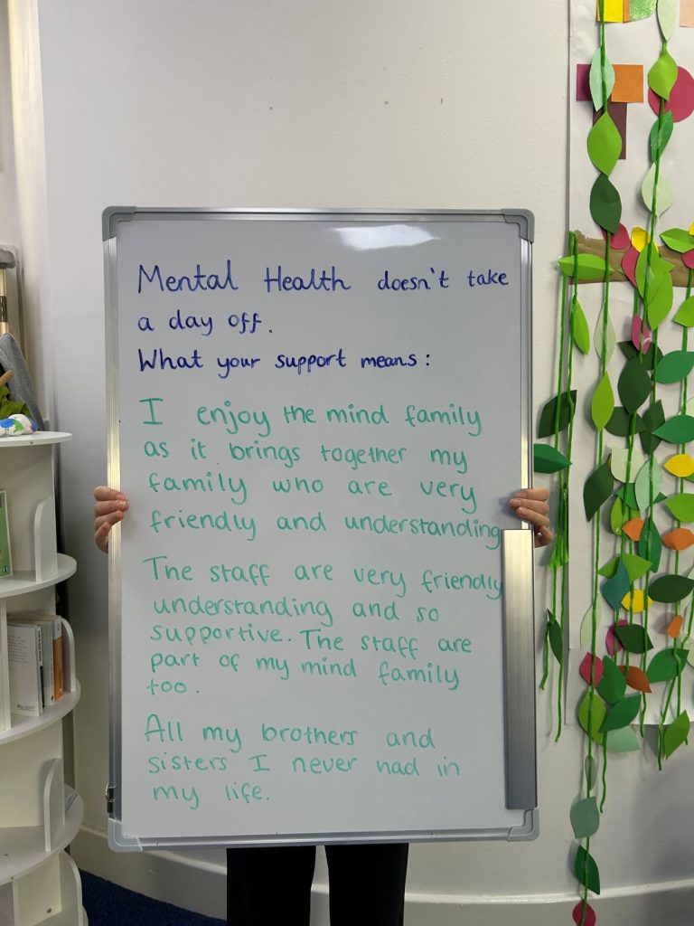 A person holds a whiteboard that says: “Mental health doesn’t take a day off. What your support means: I enjoy the Mind family as it brings together my family who are very friendly and understanding. The staff are very friendly, understanding, and supportive. The staff are part of my Mind family too. All my brothers and sisters I never had in my life.”