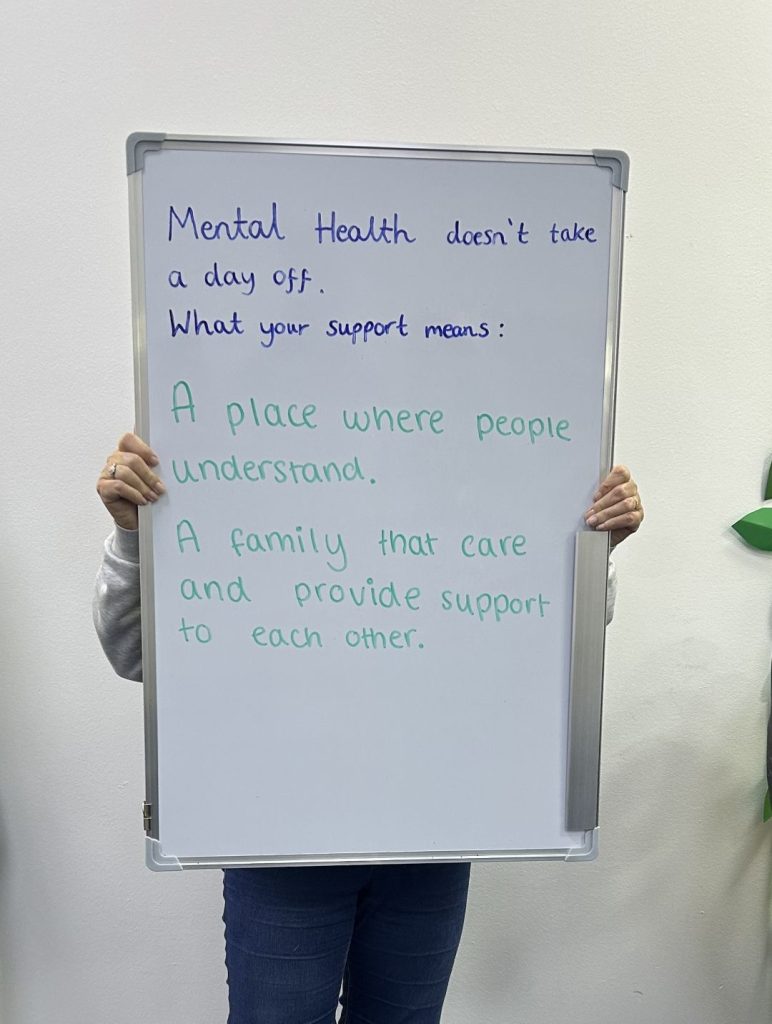A person holds a whiteboard that reads: “Mental health doesn’t take a day off. What your support means: A place where people understand. A family that care and provide support to each other.”