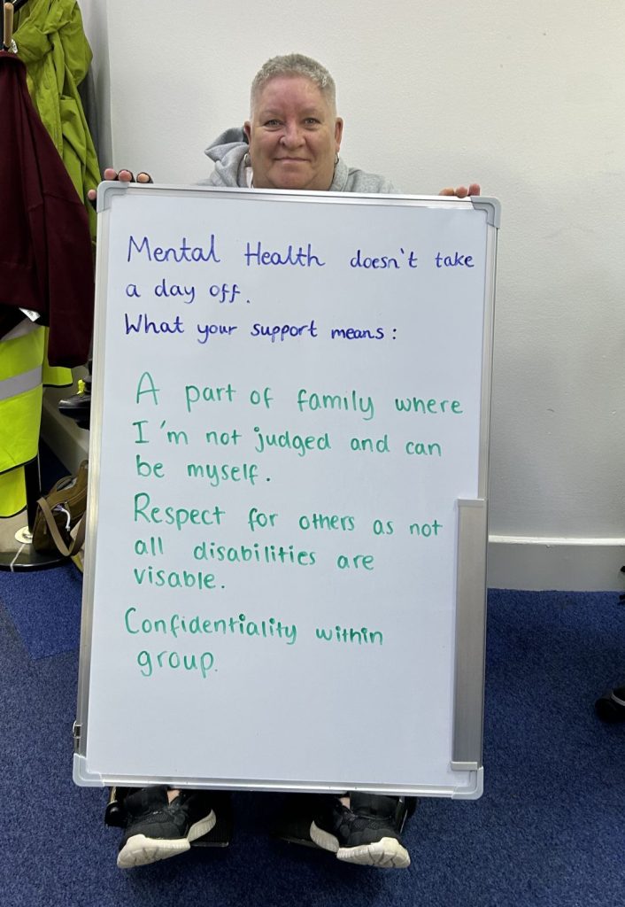 A smiling person sitting on the floor holds a whiteboard that says: “Mental health doesn’t take a day off. What your support means: A part of family where I’m not judged and can be myself. Respect for others as not all disabilities are visible. Confidentiality within group.”