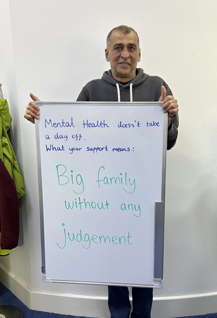 A person in a grey hoodie holds a whiteboard that says: “Mental health doesn’t take a day off. What your support means: Big family without any judgement.”