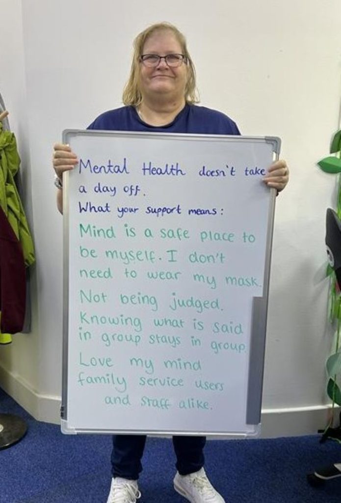 A smiling person wearing glasses and a blue top holds a whiteboard that says: “Mental health doesn’t take a day off. What your support means: Mind is a safe place to be myself. I don’t need to wear my mask. Not being judged. Knowing what is said in group stays in group. Love my Mind family — service users and staff alike.”