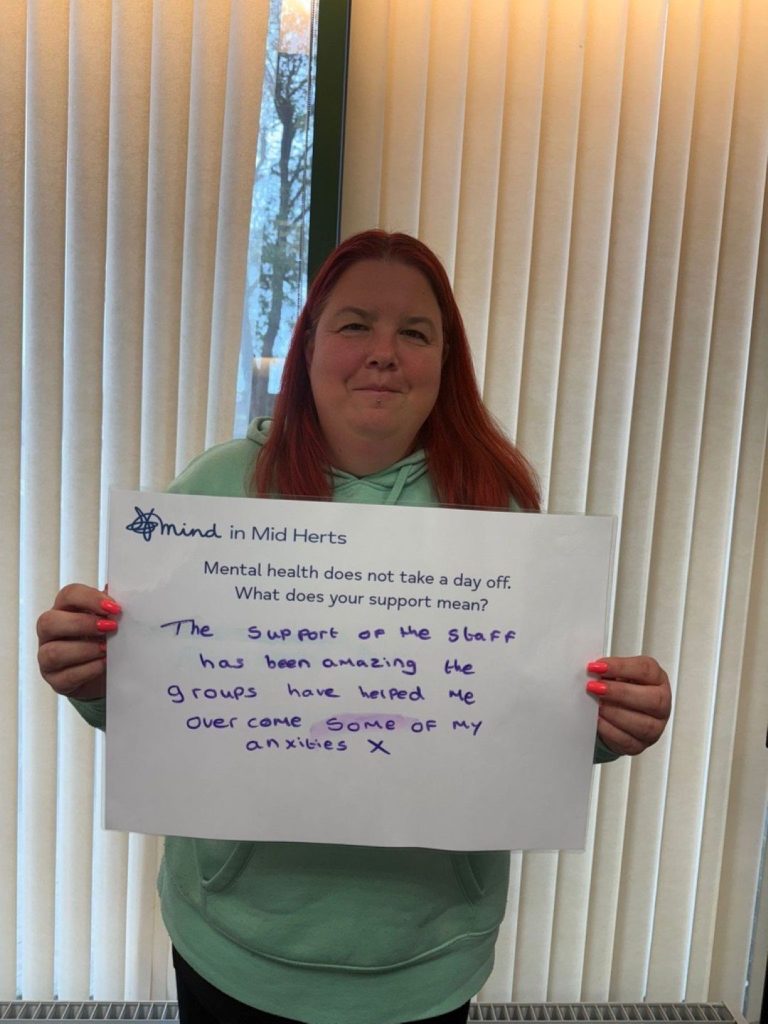A person in a light green hoodie holding a sign that says: “Mind in Mid Herts. Mental health does not take a day off. What does your support mean? The support of the staff has been amazing. The groups have helped me overcome some of my anxieties.”