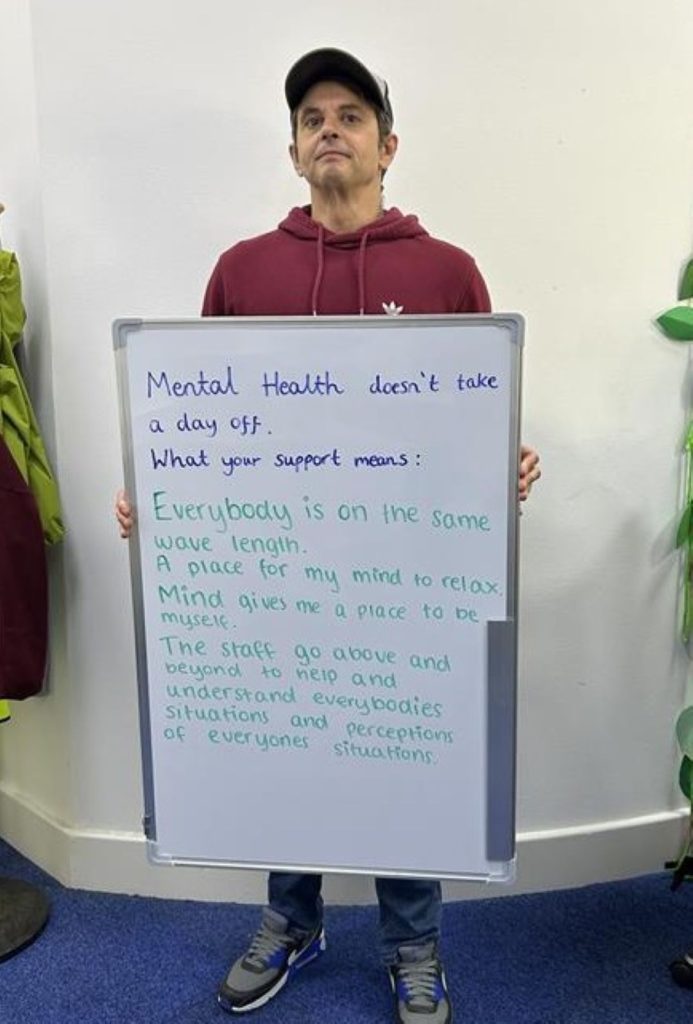 A person wearing a burgundy hoodie and cap holds a whiteboard that says: “Mental health doesn’t take a day off. What your support means: Everybody is on the same wavelength. A place for my mind to relax. Mind gives me a place to be myself. The staff go above and beyond to help and understand everybody’s situations and perceptions.