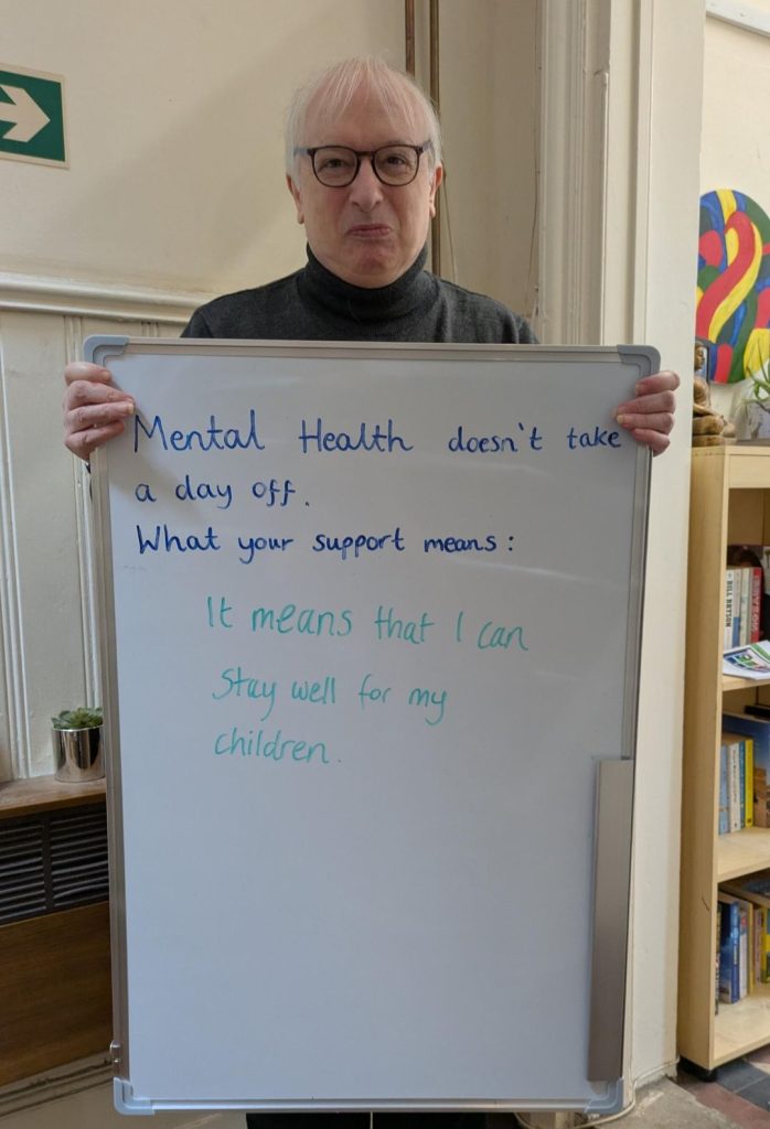 A person wearing glasses and a dark jumper holds a whiteboard that says: “Mental health doesn’t take a day off. What your support means: It means that I can stay well for my children.”