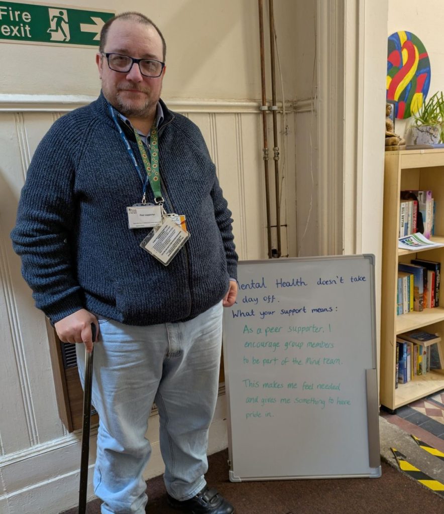 A person wearing glasses and a lanyard with ID badges stands next to a whiteboard that reads: “Mental health doesn’t take a day off. What your support means: As a peer supporter, I encourage group members to be part of the Mind team. This makes me feel needed and gives me something to have pride in.”