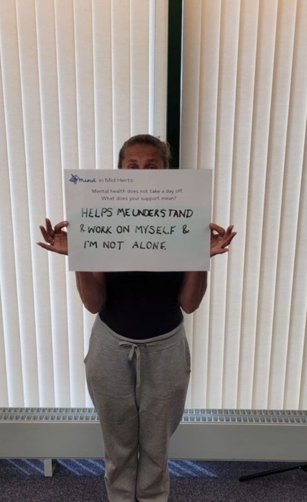 A person holding a sign that says: “Mind in Mid Herts. Mental health does not take a day off. What does your support mean? Helps me understand and work on myself and I’m not alone.”