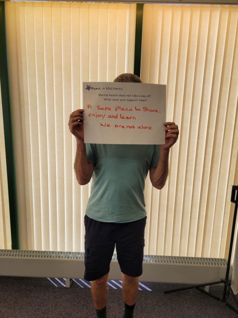 A person holding a sign that says: “Mind in Mid Herts. Mental health does not take a day off. What does your support mean? A safe place to share, enjoy and learn. We are not alone.”