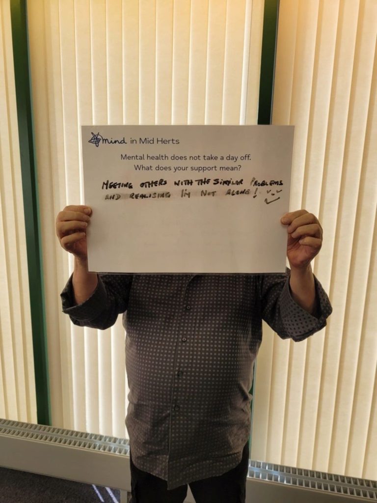 A person holding a sign that says: “Mind in Mid Herts. Mental health does not take a day off. What does your support mean? Meeting others with similar problems and realising I’m not alone.”