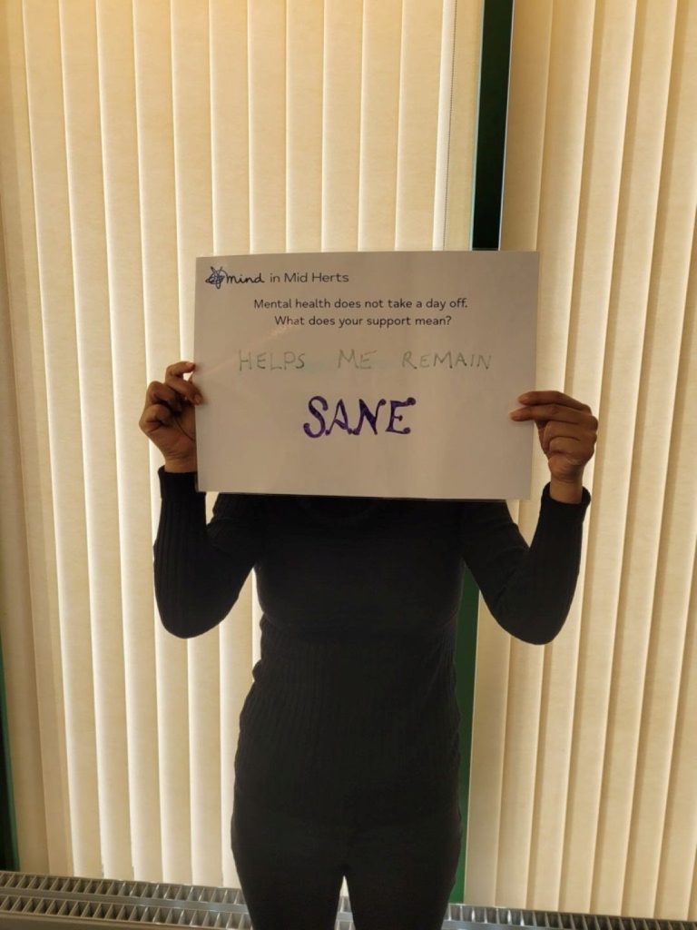 A person standing in front of cream blinds, holding a sign that reads: “Mind in Mid Herts. Mental health does not take a day off. What does your support mean? Helps me remain sane.”
