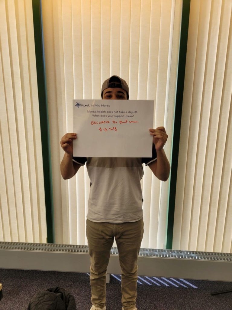A person wearing a cap and holding a sign that says: “Mind in Mid Herts. Mental health does not take a day off. What does your support mean? Becoming the best version of myself.”