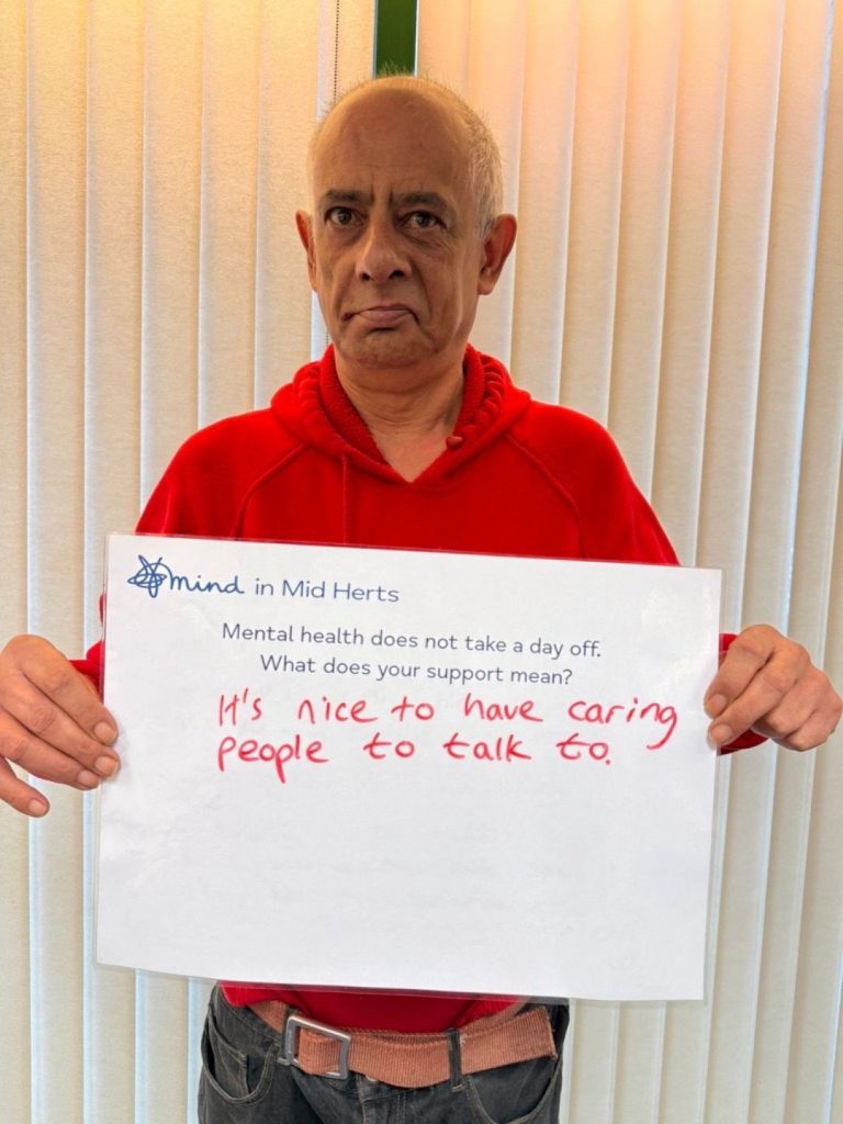 A person in a red jumper holding a sign that says: “Mind in Mid Herts. Mental health does not take a day off. What does your support mean? It’s nice to have caring people to talk to.”