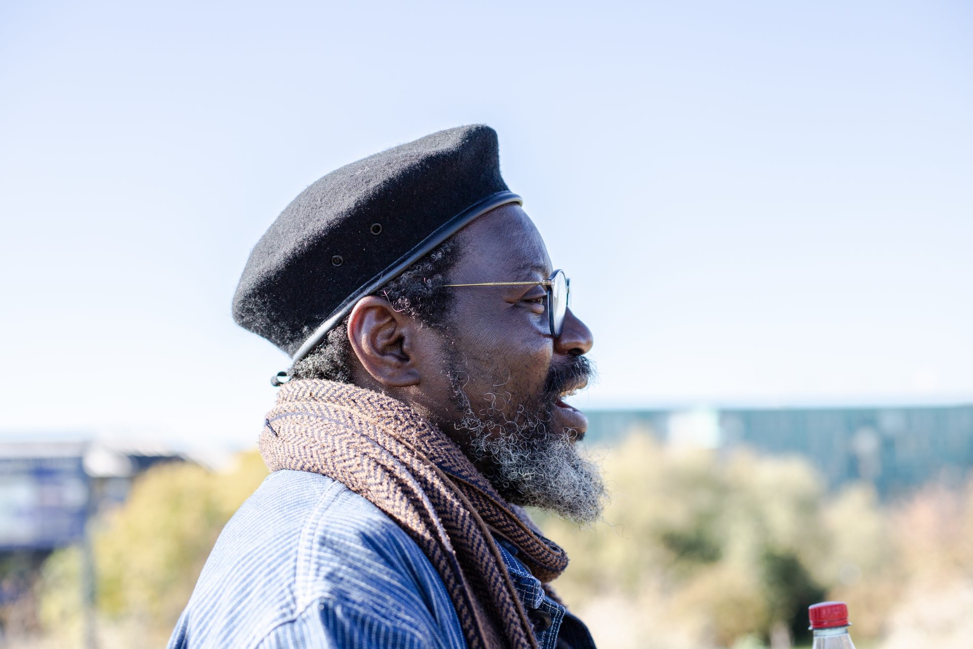 A man with a greying beard wearing glasses, a black beret, and a brown scarf is pictured in profile outdoors on a sunny day. The background is softly blurred, showing trees and buildings under a clear blue sky.