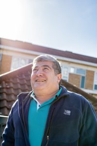 A man smiles warmly while standing outdoors on a sunny day, with a tiled roof and building in the background. He is wearing a teal polo shirt under a navy jacket.